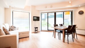 Uma sala de estar moderna com sofá, mesa de jantar com cadeiras, televisão de tela plana e janelas amplas.
