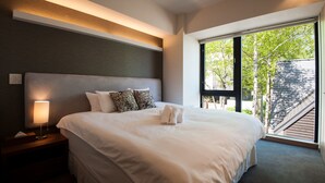 Quarto de hotel moderno com uma cama grande, uma mesa de cabeceira com abajur e vista para o exterior através de uma janela ampla.
