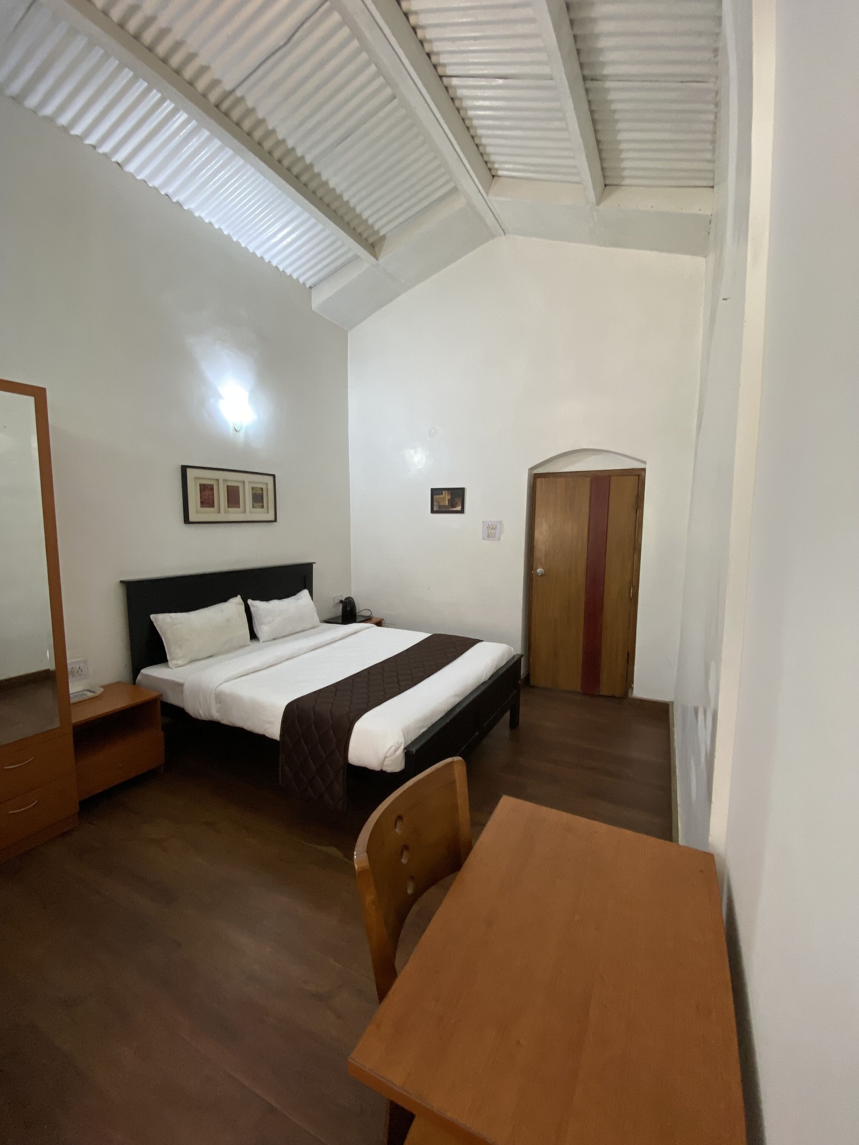 A bedroom with a bed, a wooden nightstand, a wooden chair, a wooden table, a mirror, and a door.