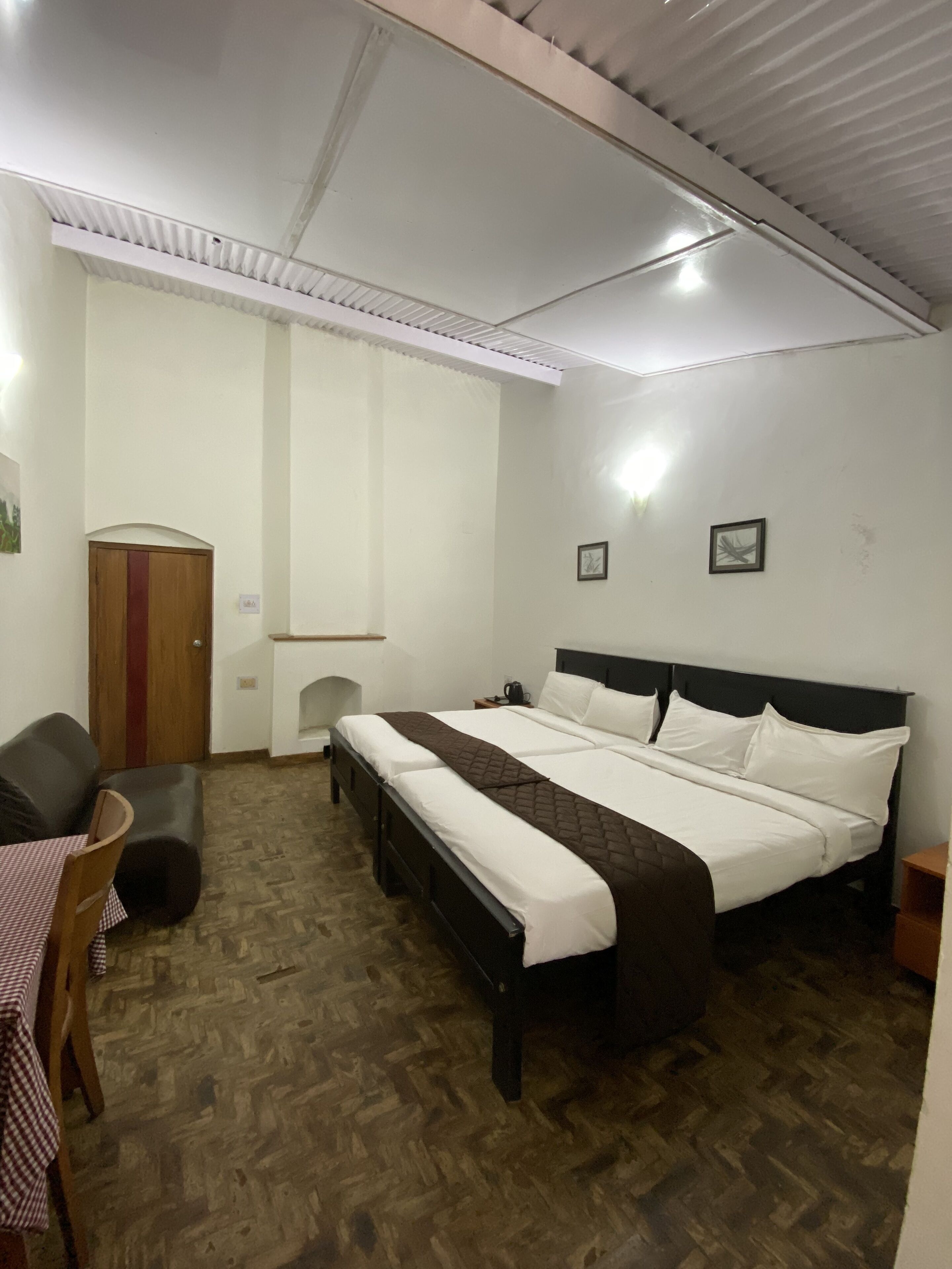 A room with a large bed, a wooden wardrobe, a small table with a checkered tablecloth, and a chair.