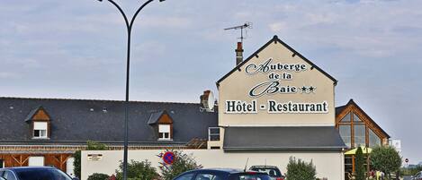 Un hotel llamado Auberge de la Baie, con un letrero que indica que es un hotel y restaurante. Hay coches estacionados adelante y se ve una farola.