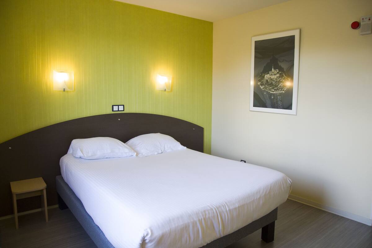 Una cama bien hecha en una habitación con paredes verdes, dos luces fijadas a la pared y un cuadro enmarcado.