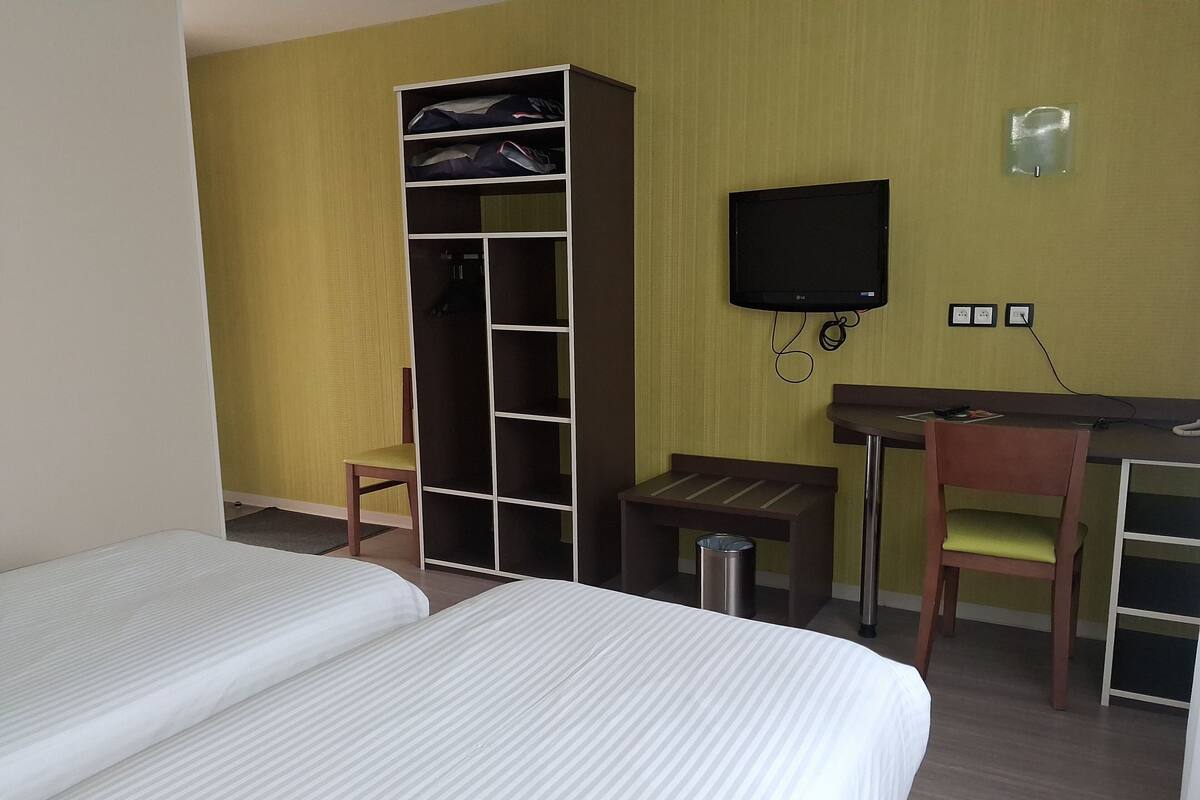 Habitación de hotel con dos camas, un armario, un escritorio, una silla, un televisor y un cubo de basura.