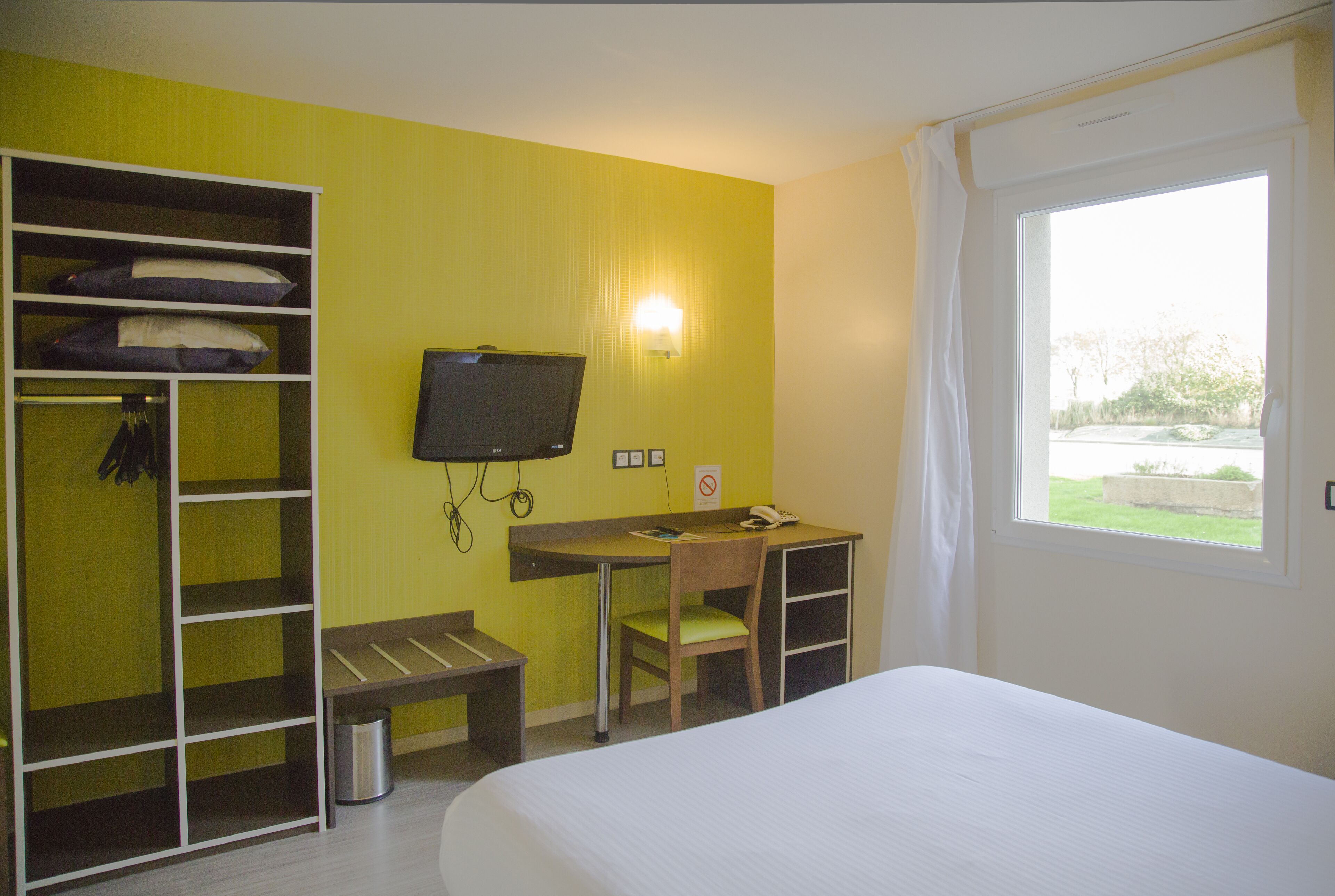 A hotel room with a bed, a desk with a chair, a television, a wardrobe, and a window with a view of greenery.