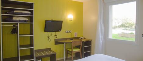 A hotel room with a bed, a desk with a chair, a television, a wardrobe, and a window with a view of greenery.