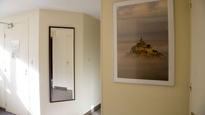A hallway with a framed picture of a castle on a cloudy day, a mirror on the left, and a door at the end. - Auberge de la Baie (Pontorson)