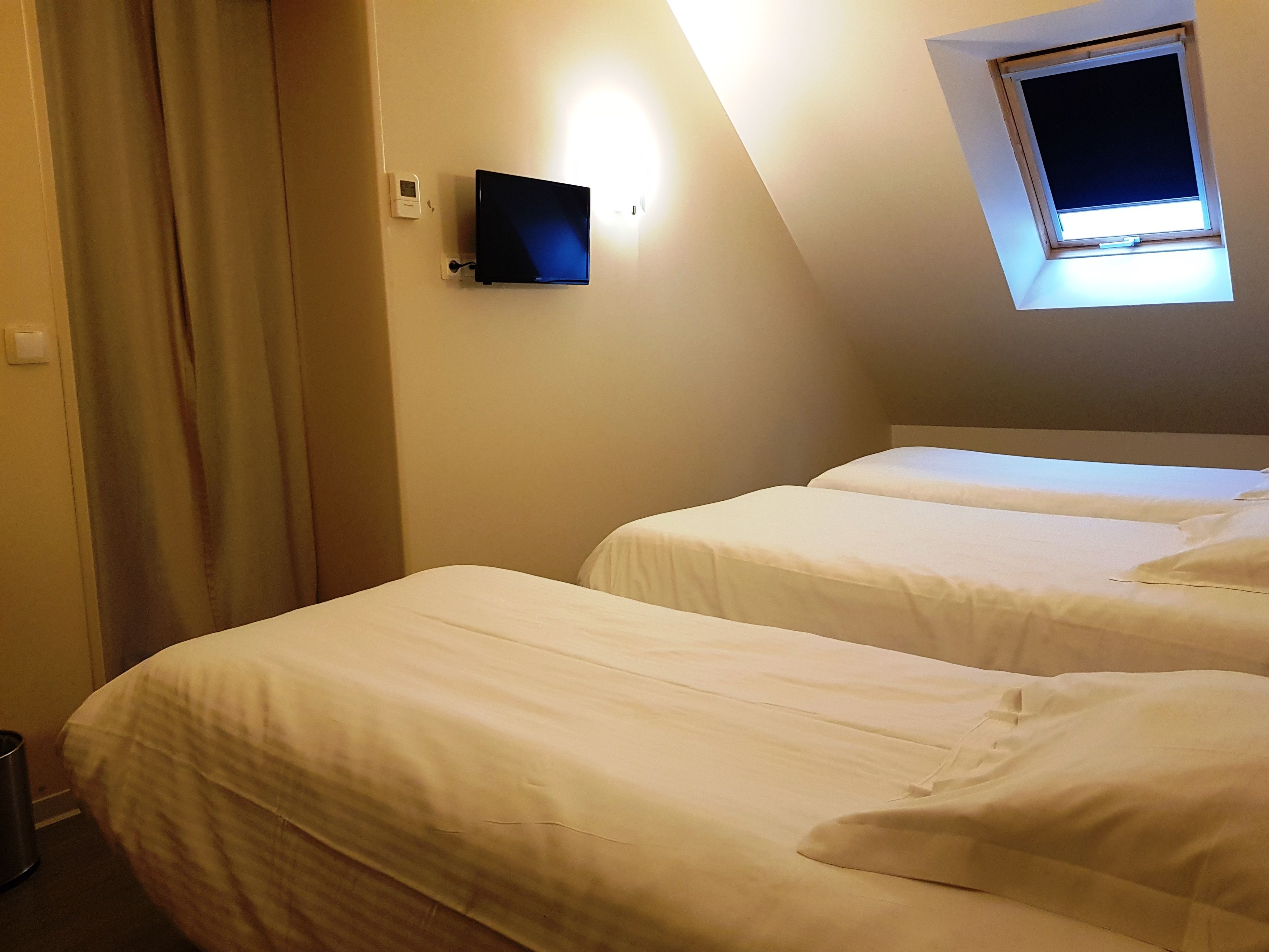 A hotel room with two beds, a TV mounted on the wall, and a window with a view of the sky.