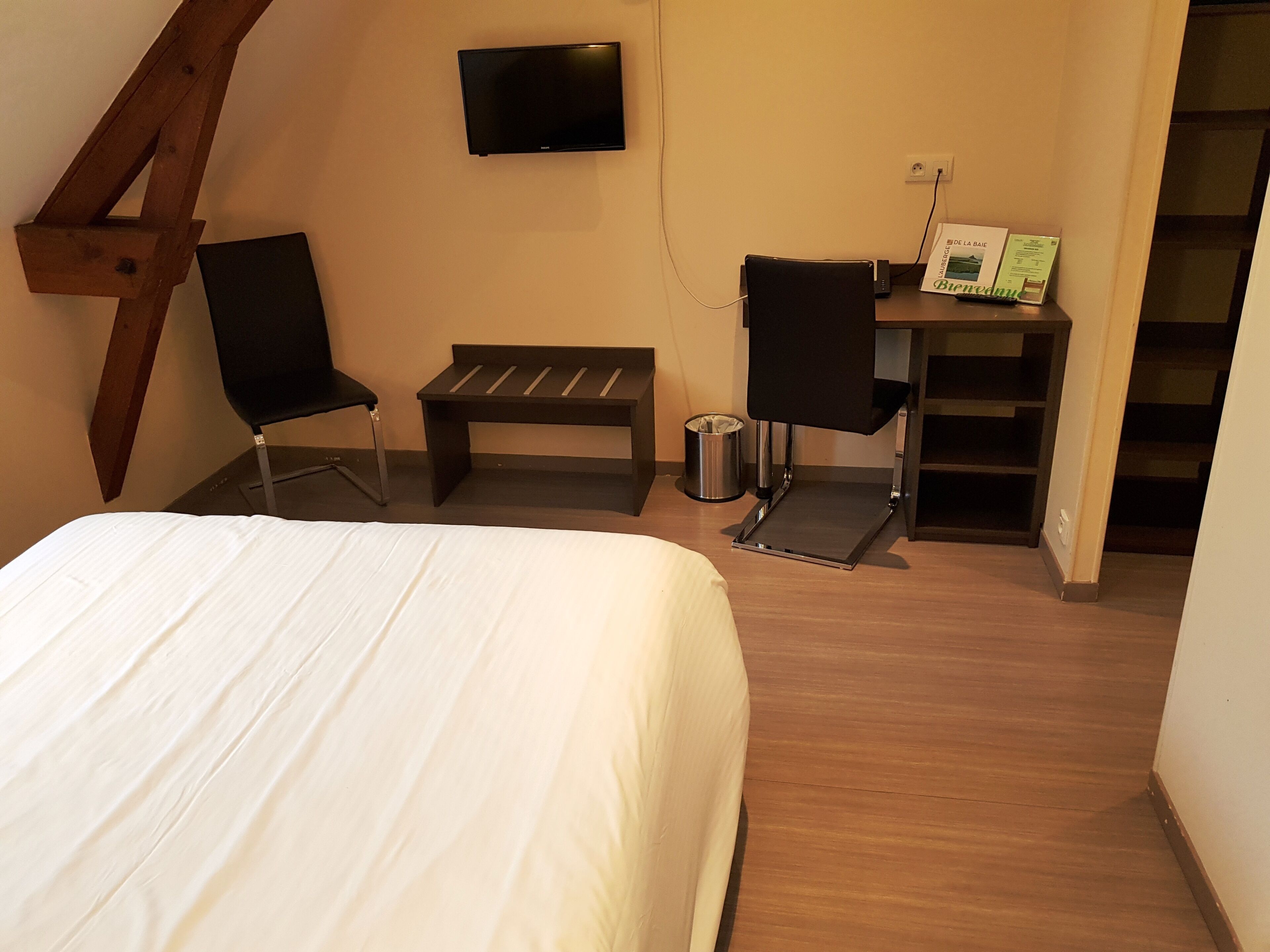 Quarto com cama, escrivaninha com cadeira, um banquinho e televisão na parede.