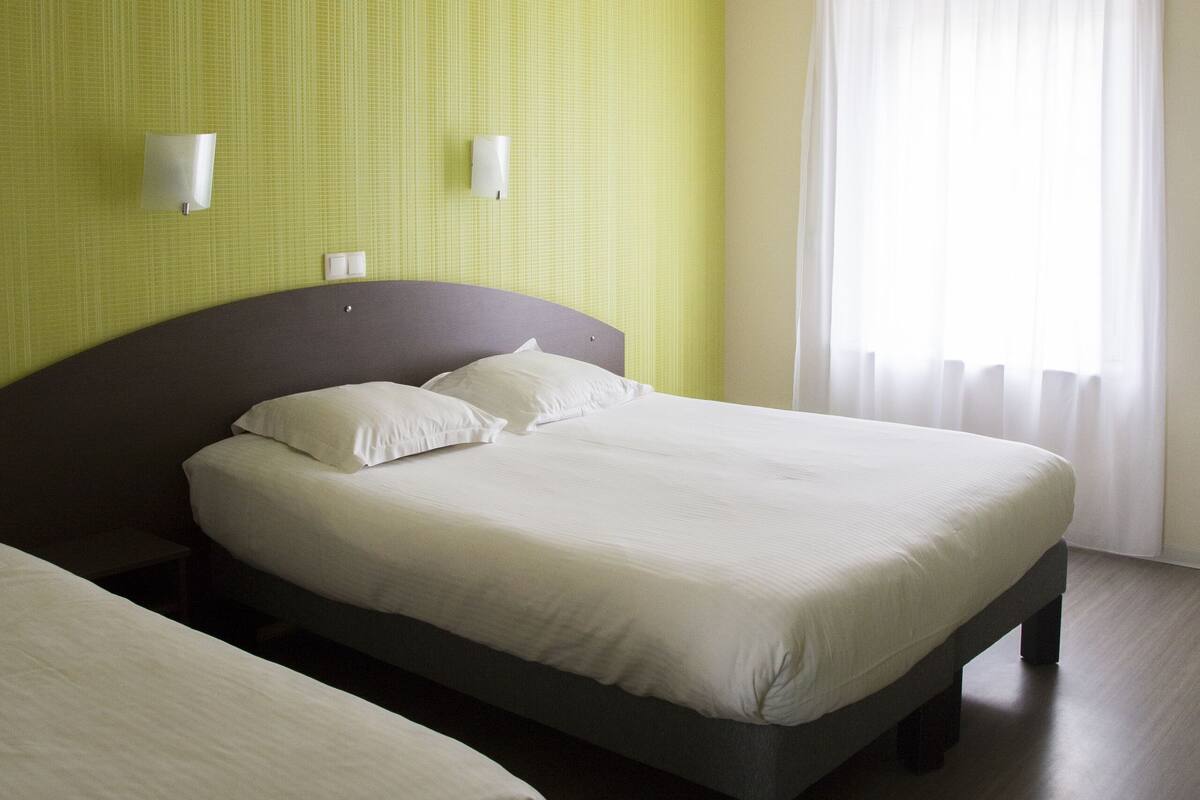 Una habitación de hotel con dos camas, un ventanal con cortinas traslúcidas y lámparas fijadas a la pared.