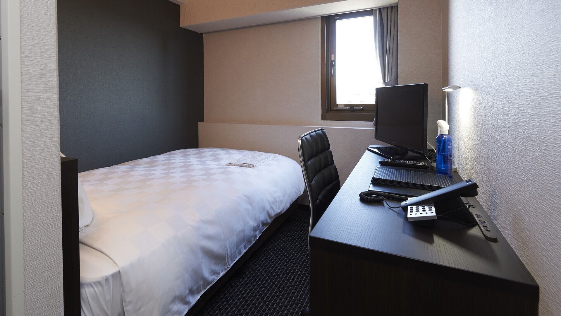 A small hotel room with a single bed, a desk with a computer, and a window.