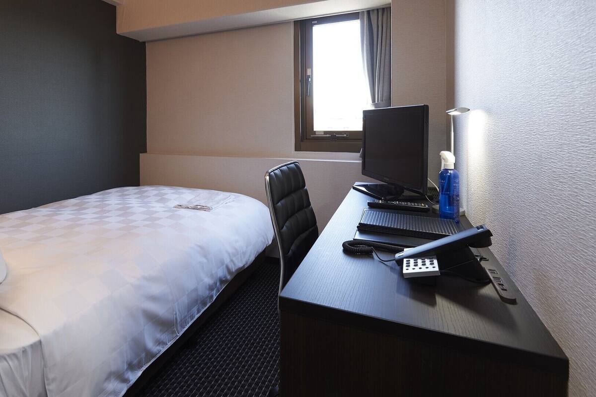 Pieni hotellihuone, jossa on yksi sänky, työpöytä tietokoneella ja ikkuna.