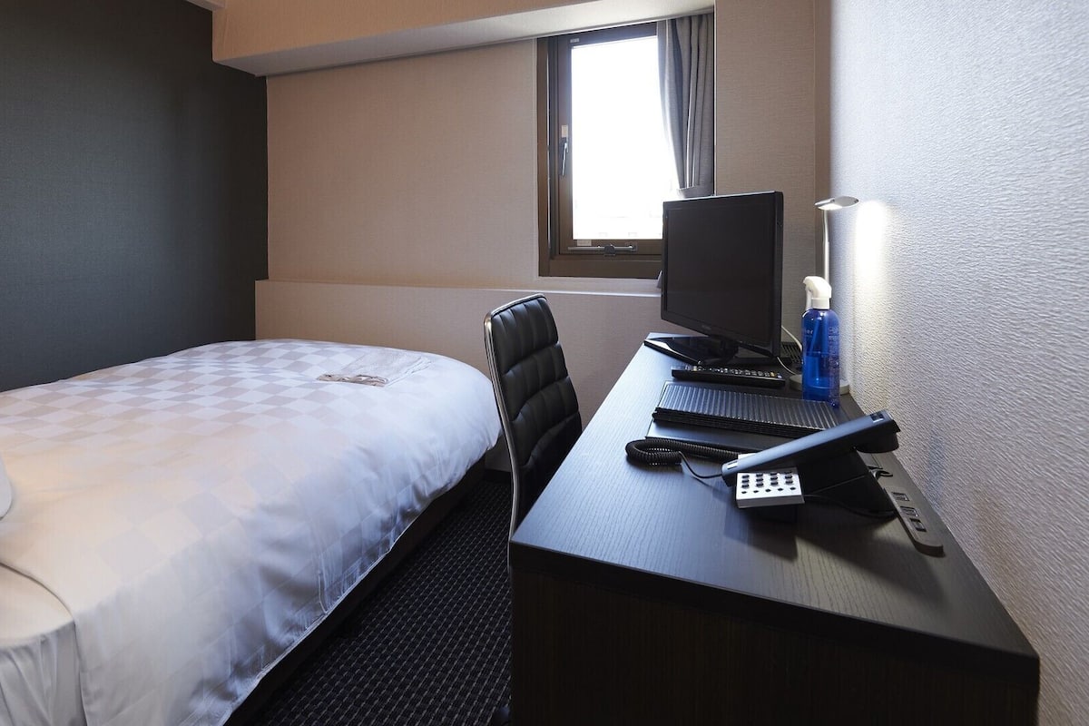 A small hotel room with a single bed, a desk with a computer, and a window.