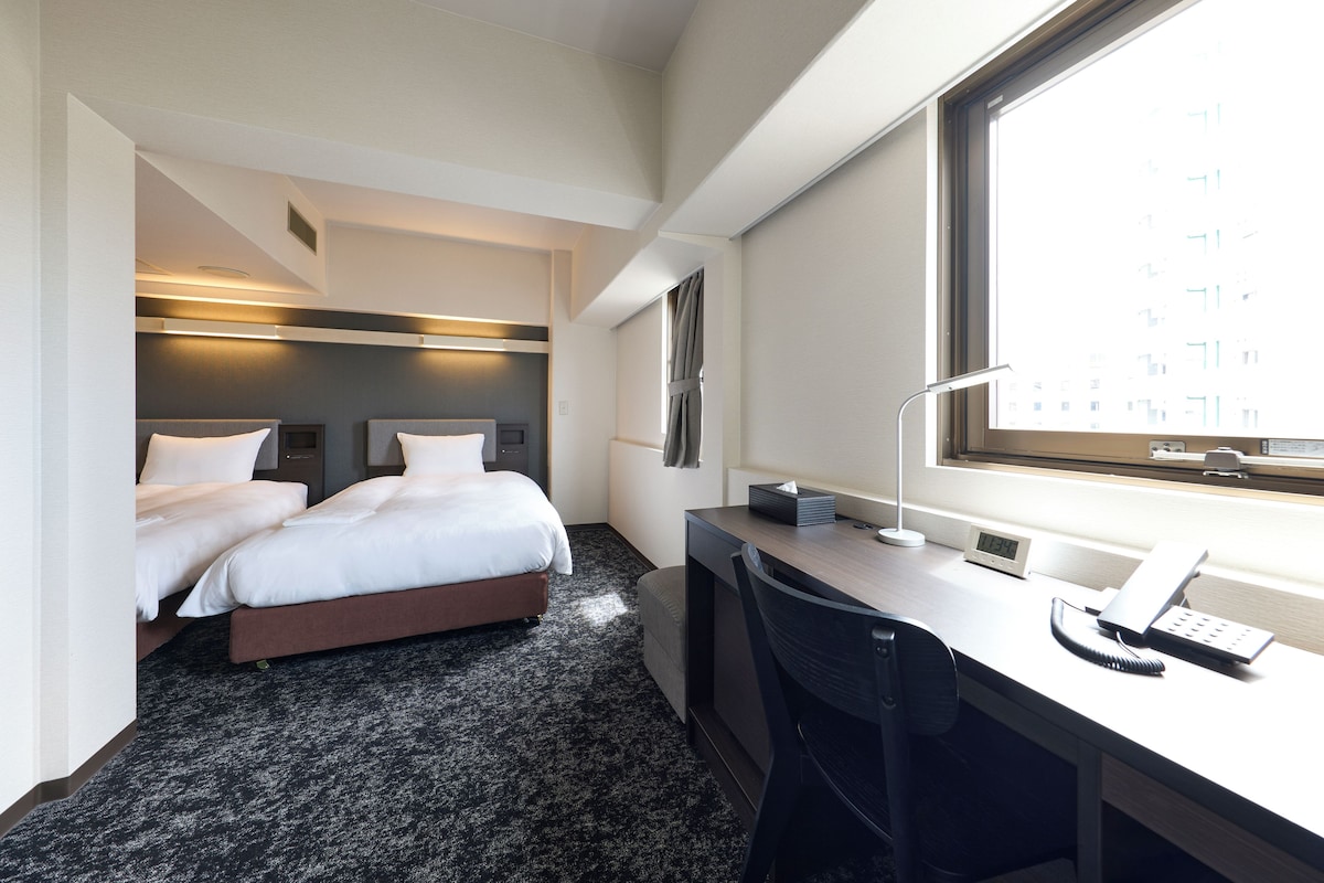 Habitación de hotel con dos camas, un escritorio, una silla, un ventanal y un reloj montado en la pared.