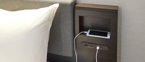 A bed with a pillow, a bedside table with a phone charging, and a power outlet.