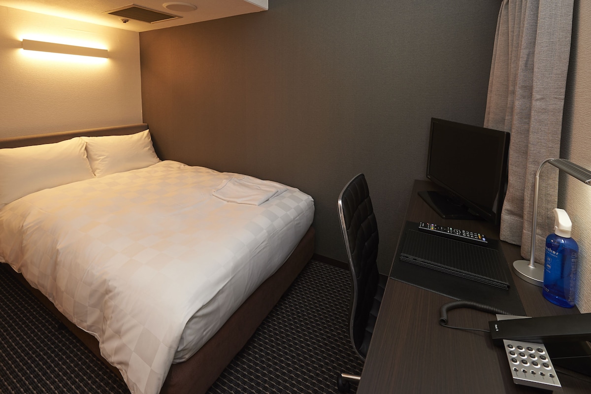 Una habitación de hotel con cama, escritorio, computadora, teléfono y un frasco de gel antibacterial.