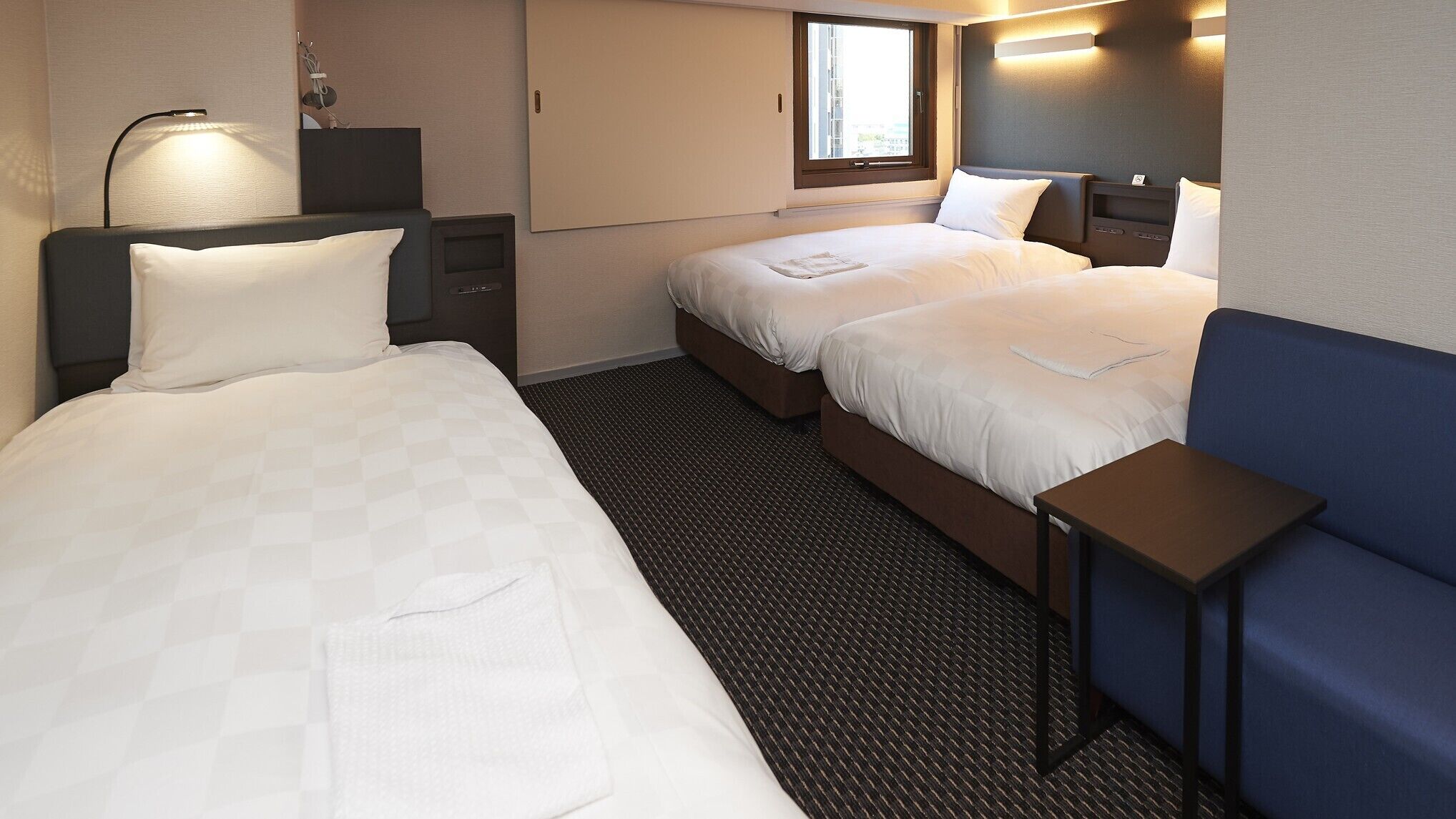 A hotel room with two beds, a desk, a chair, a lamp, and a window.