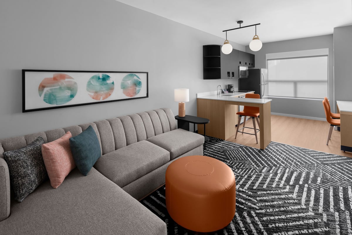 A modern living room with a grey sectional sofa, a round orange ottoman, and a black and white patterned rug. There is a kitchen area with a bar counter and orange chairs.