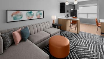 A modern living room with a grey sectional sofa, a round orange ottoman, and a black and white patterned rug. There is a kitchen area with a bar counter and orange chairs.
