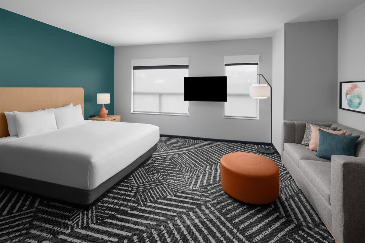 A hotel room with a large bed, a sofa, a small orange ottoman, two windows, a flat-screen TV, and a nightstand with a lamp.