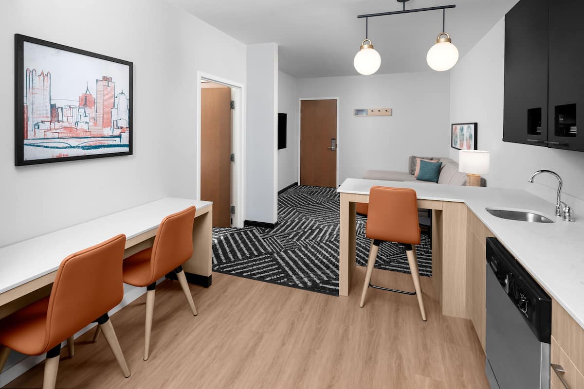 A modern kitchen with a dishwasher, a dining area with two chairs, a black and white patterned rug, and a cityscape painting on the wall.