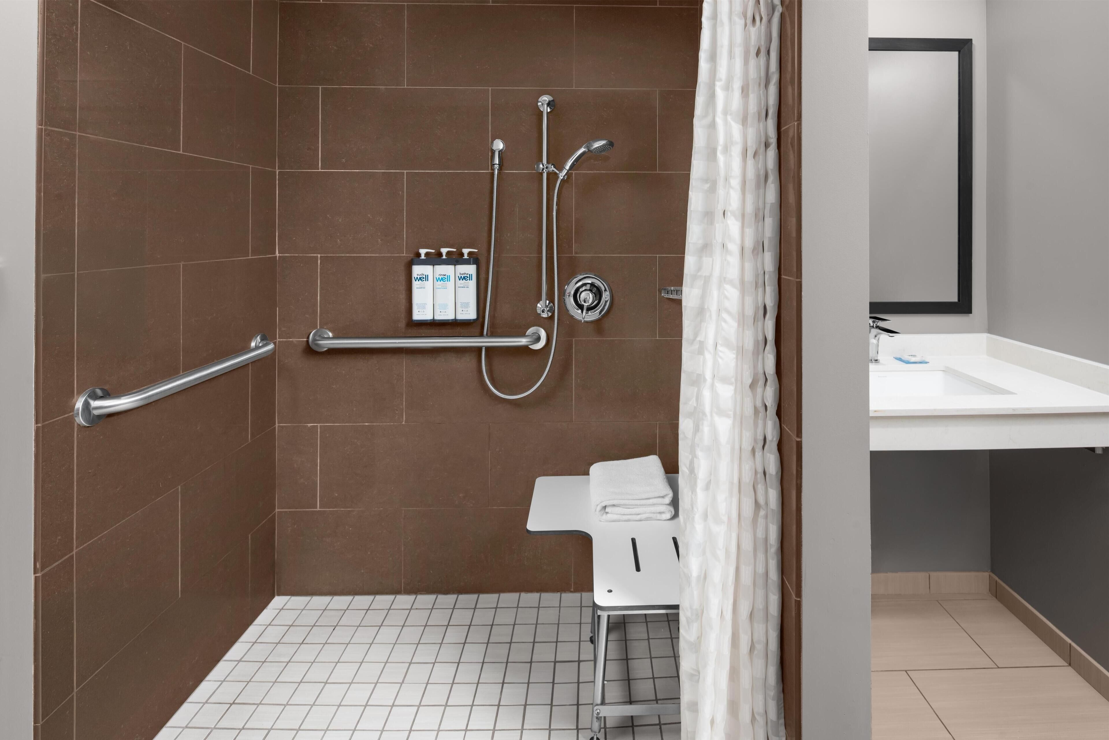 Un baño moderno con baldosas marrones, un lavamanos blanco, un espejo y una zona de ducha con barras de apoyo y cabezal de ducha manual.