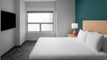 A hotel room with a large bed, a television on the wall, a window, and a nightstand with a lamp.