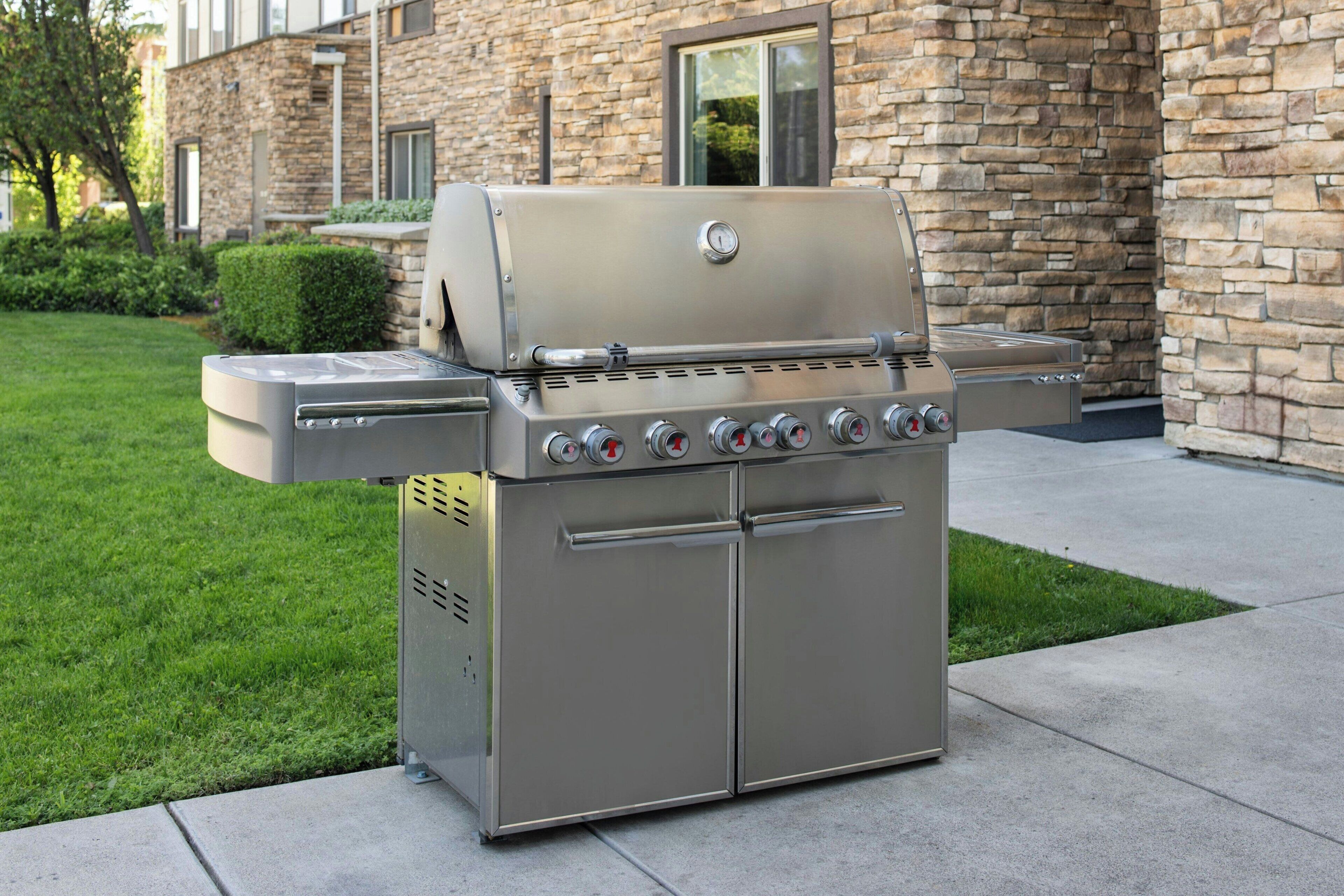 A stainless steel outdoor grill with a digital thermometer, located in a landscaped area with a stone building in the background.