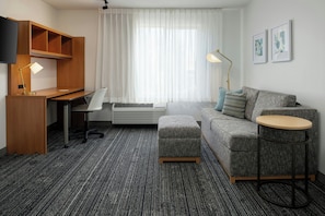 A hotel room with a desk, chair, sofa, ottoman, round table, and artwork on the wall.