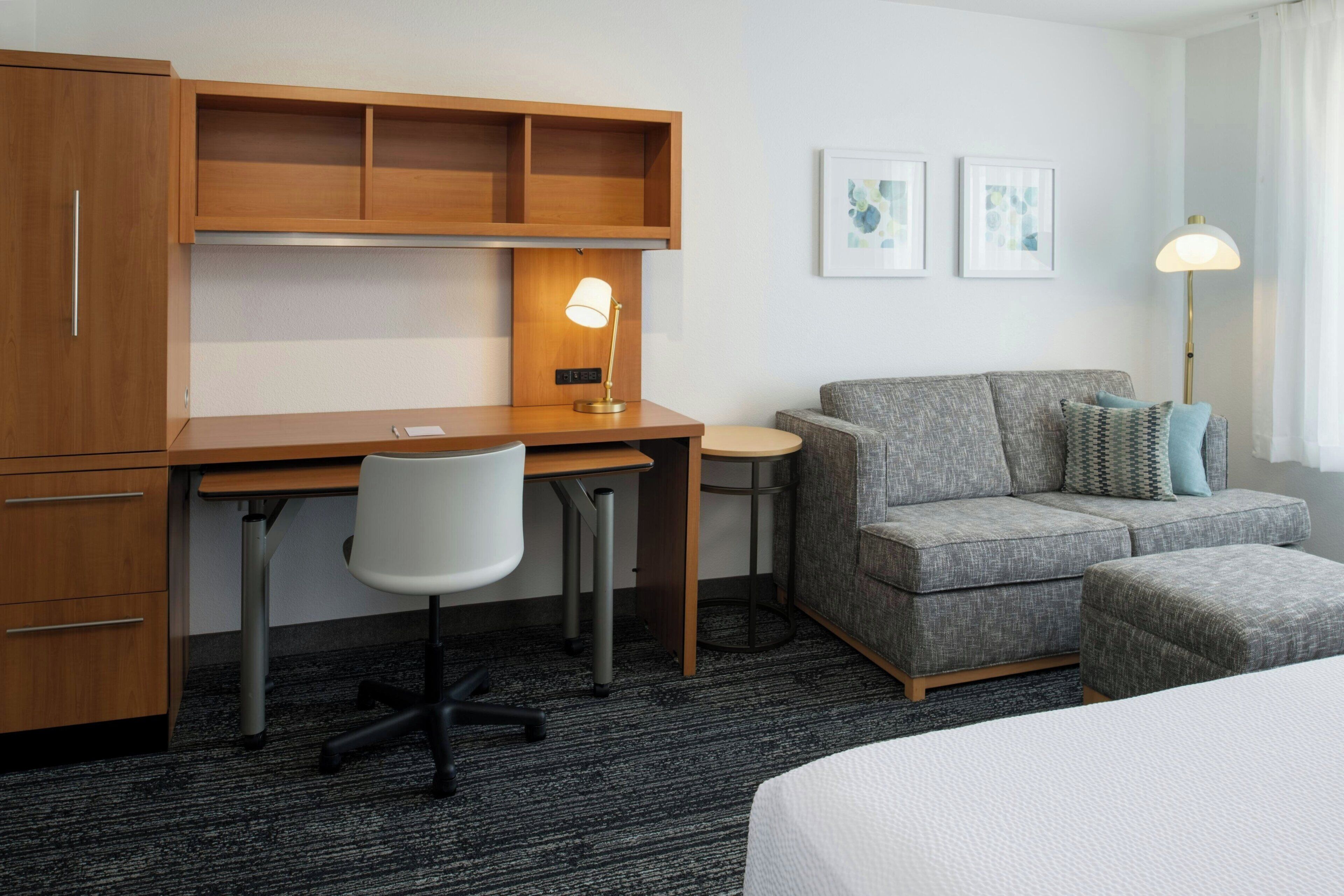 A hotel room with a wooden desk, a gray sofa, a small round table, a white chair, and a bed.