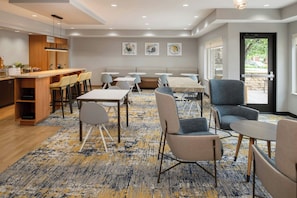 A modern hotel room with a kitchen area, a dining area with tables and chairs, and a large window with a view.