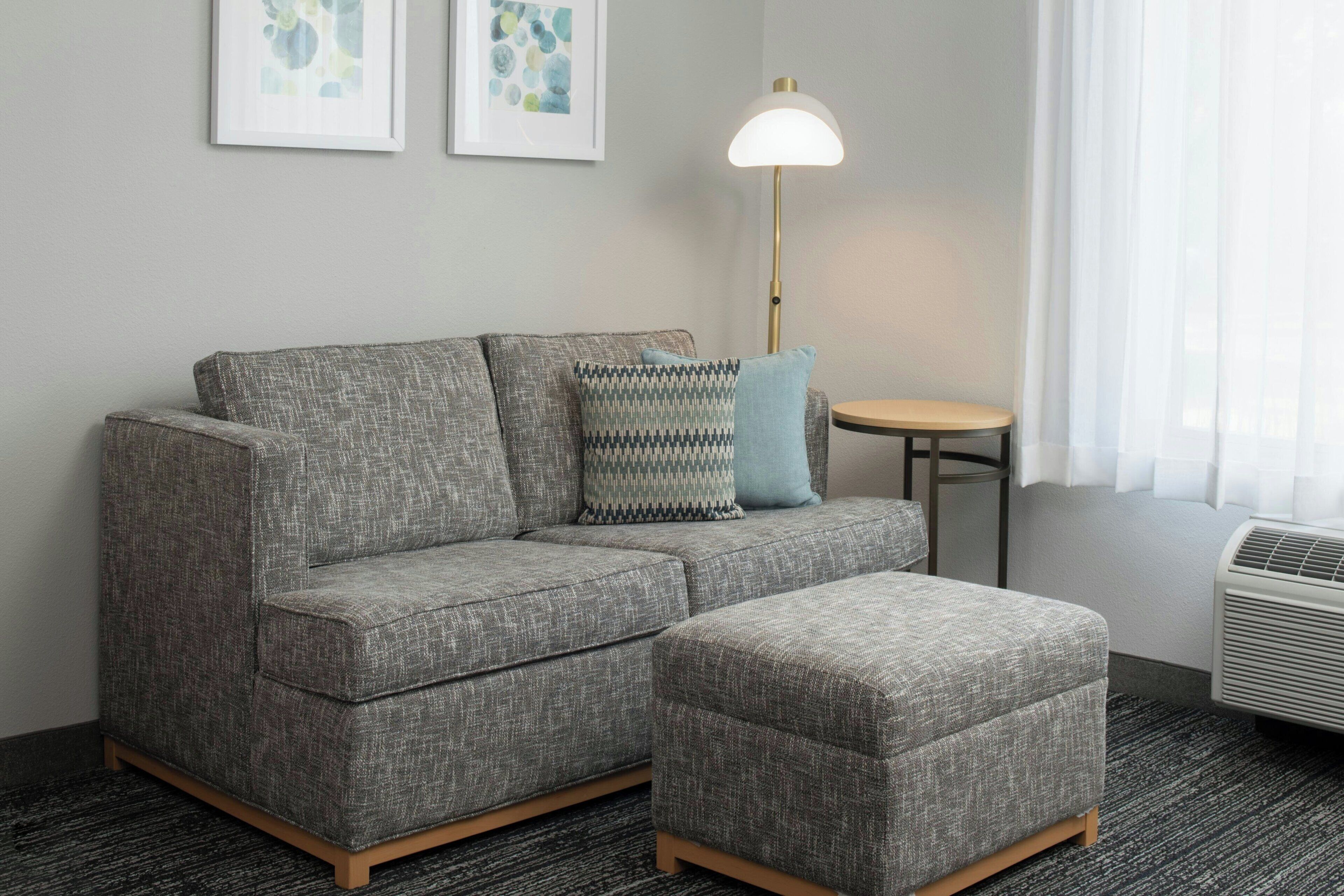A gray sectional sofa with a matching ottoman, a small round table, a floor lamp, and two framed abstract artworks on the wall.