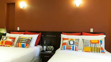A hotel room with two beds, each with colorful patterned pillows, a nightstand with a bottle and a phone, and two wall-mounted lights.