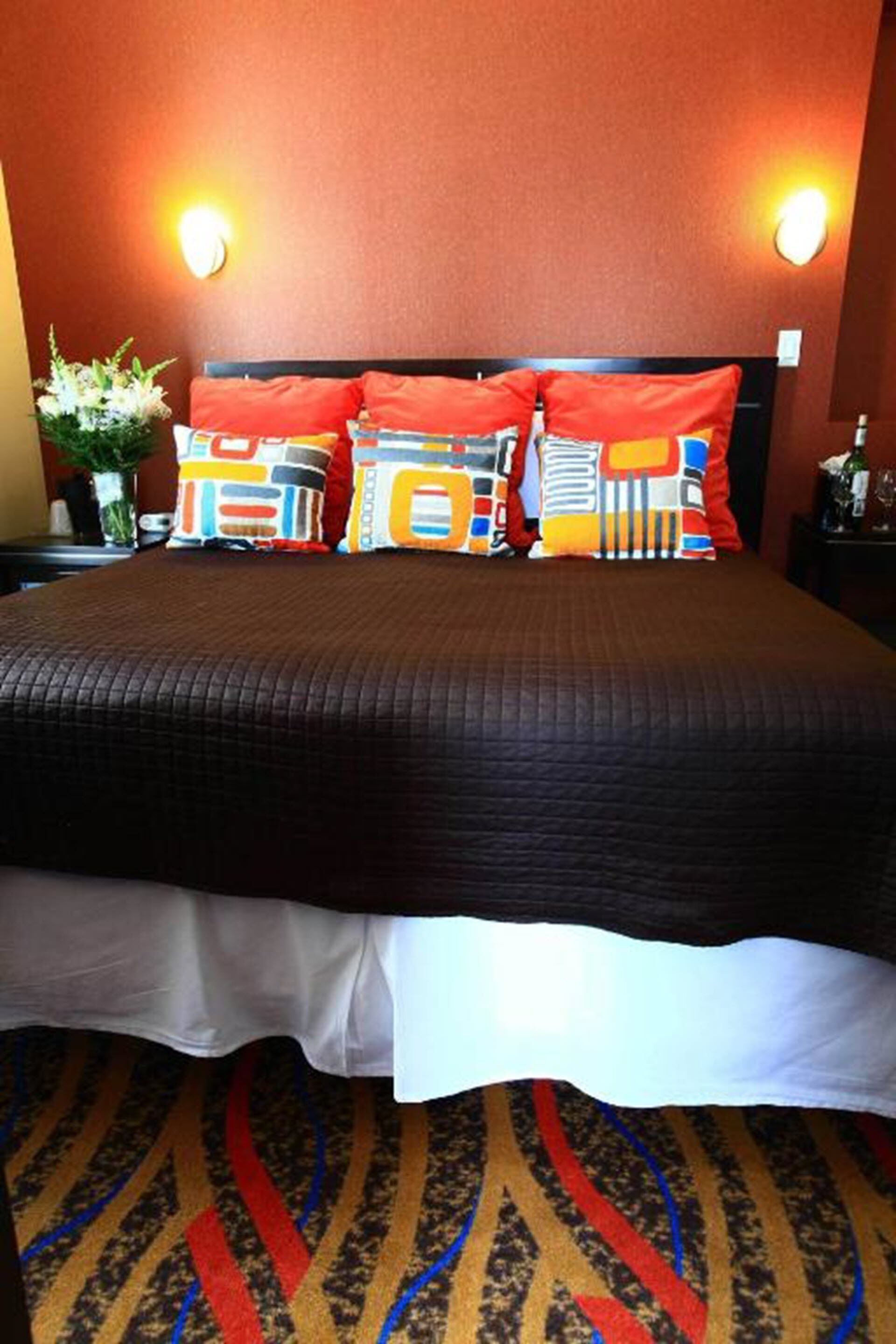 A hotel room with a large bed, a headboard with multiple pillows, a nightstand with a vase of flowers, and a carpet with a striped pattern.