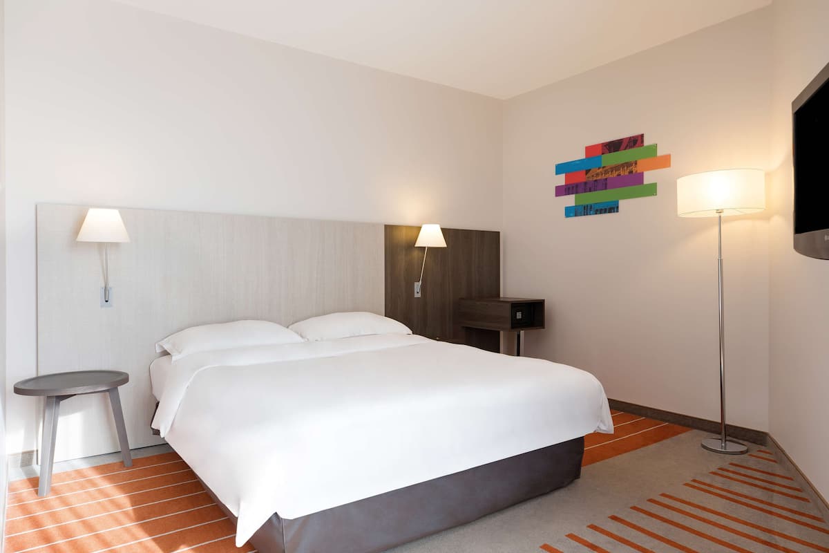 Una habitación de hotel con cama, mesita de noche, lámpara y una obra de arte en la pared.