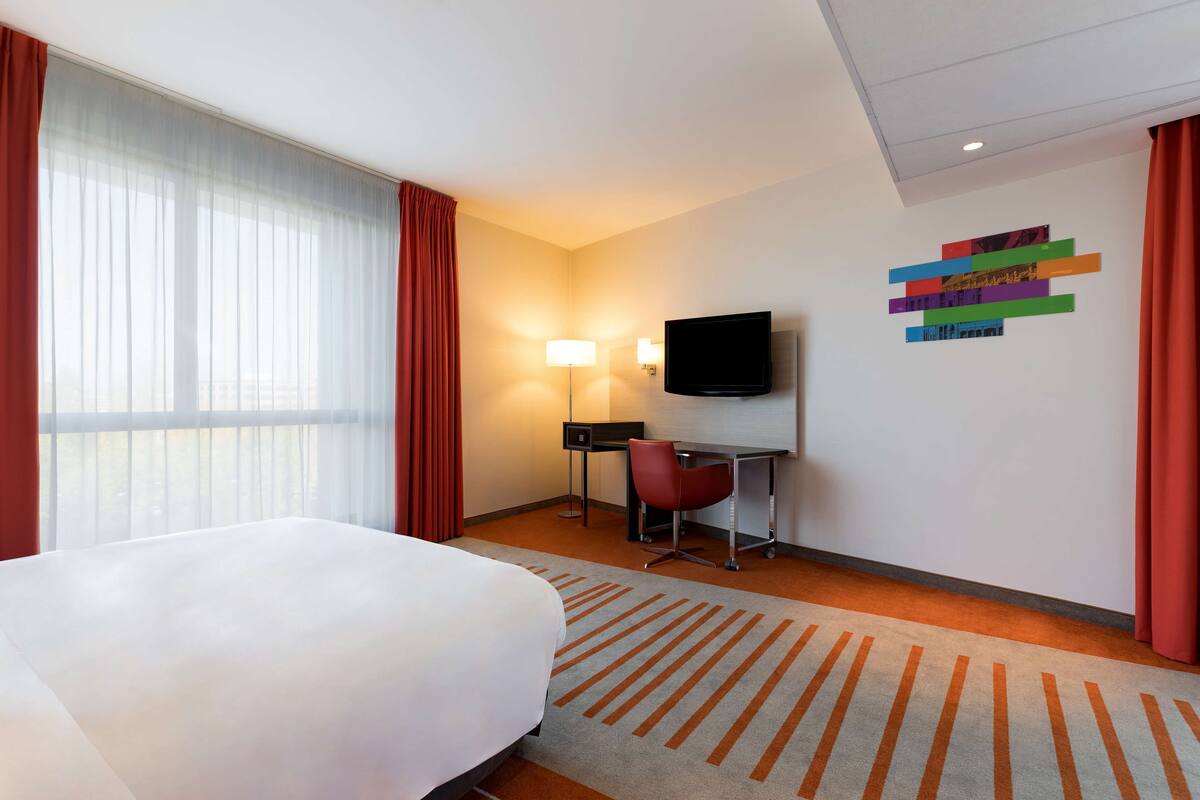 Habitación de hotel con cama, escritorio, silla, televisor y una obra de arte en la pared.