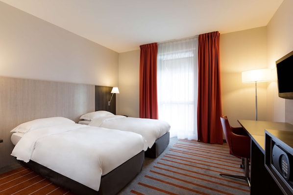 A hotel room with two beds, a desk, a TV, and a window with curtains.