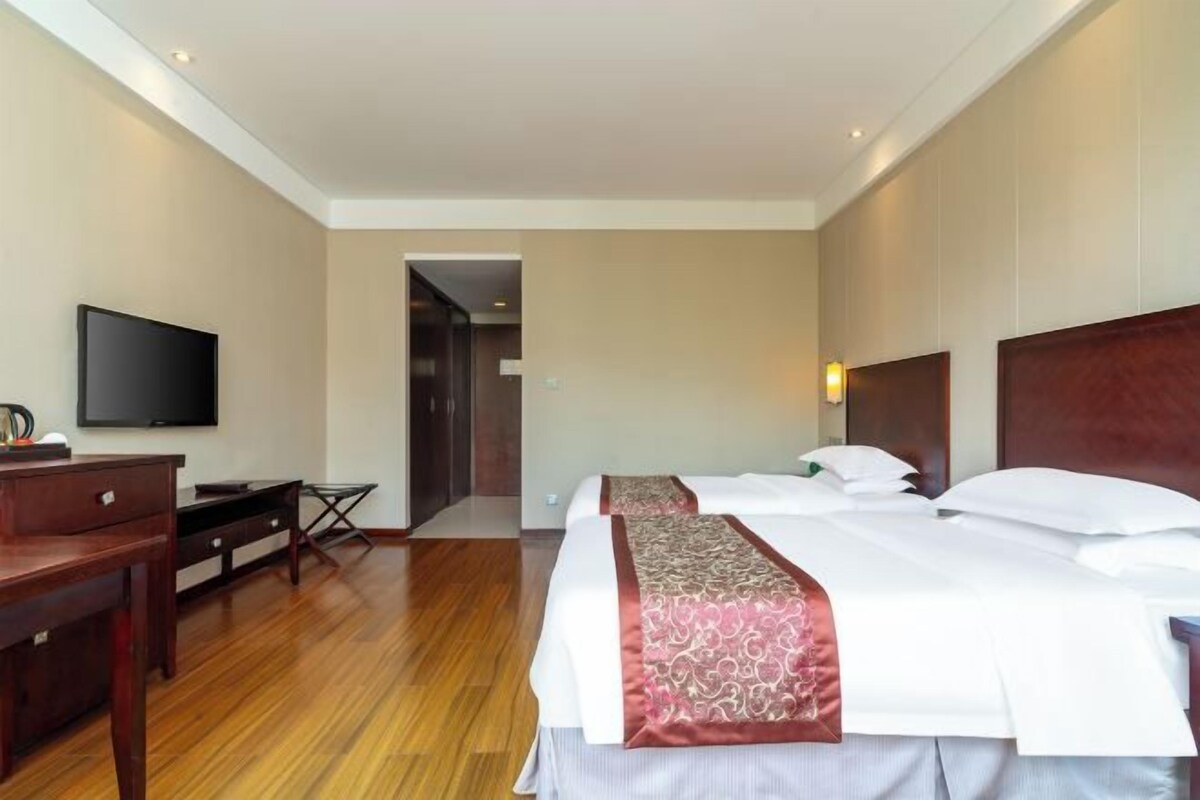 A hotel room with a wooden floor, a desk with a chair, a flat-screen TV, a bed with white bedding and a red decorative pillow, and a wall-mounted light.