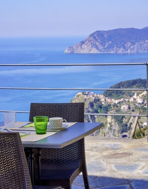 Mesa com cadeiras e um copo de vidro verde em um terraço com vista para a costa, montanhas e uma cidade ao longe.