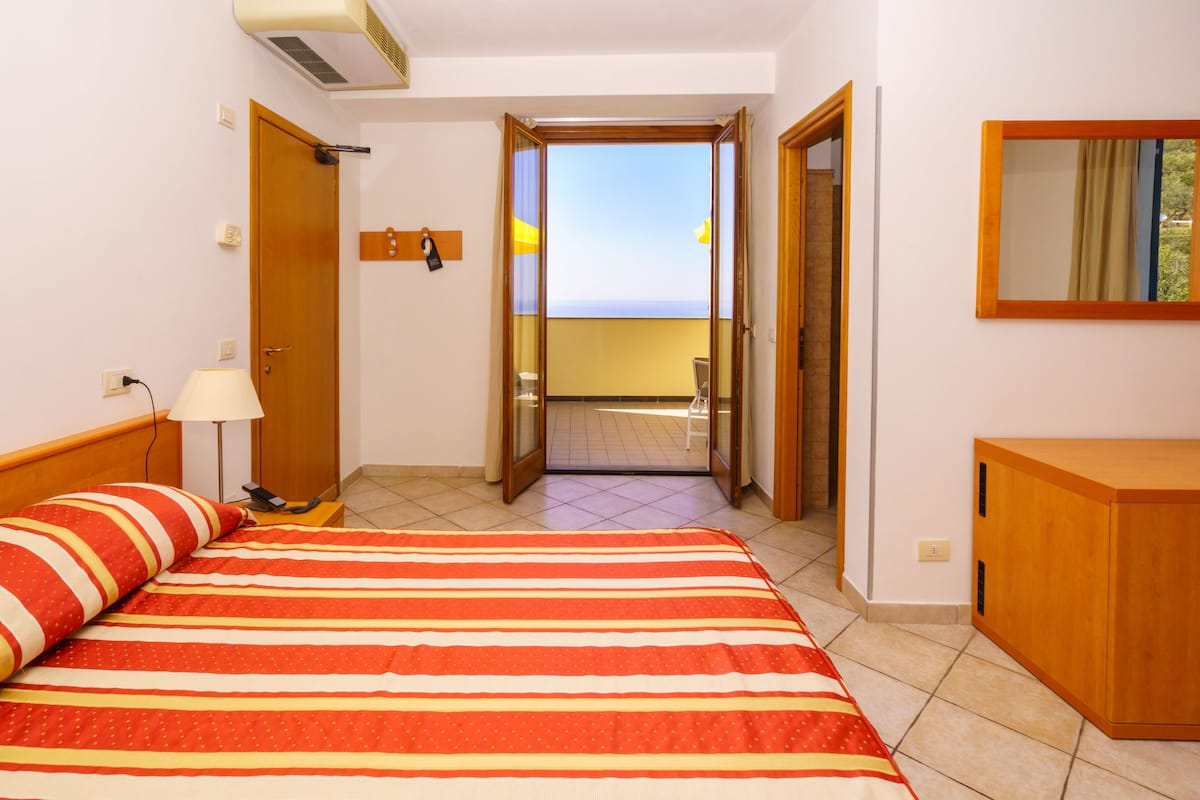 A hotel room with a bed, a nightstand, a lamp, a wooden wardrobe, and a balcony door leading to an outdoor area.