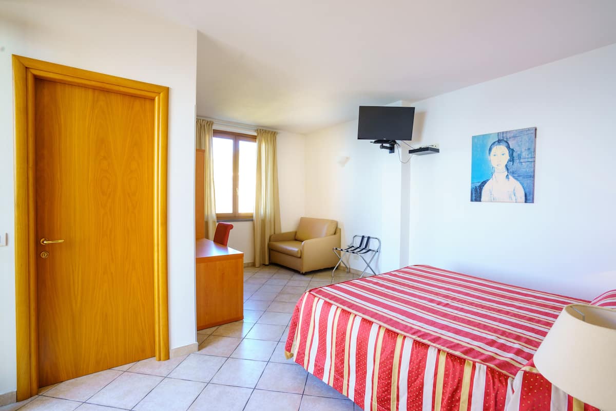 A hotel room with a wooden door, a bed with a striped bedspread, a chair, a television mounted on the wall, and a painting.