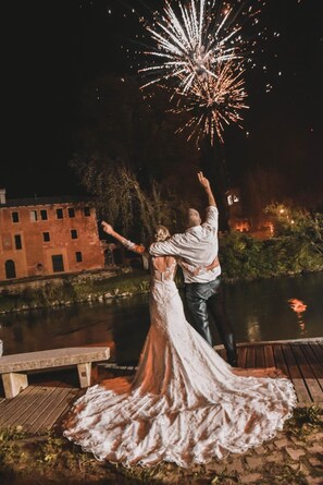 A couple embracing on a wooden dock with a river and a brick building in the background. - Principato di Ariis (Rivignano Teor)