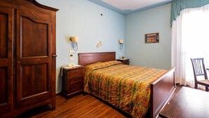A bedroom with a wooden wardrobe, a bed with a colorful bedspread, a nightstand with a lamp, and a chair by the window. - Principato di Ariis (Rivignano Teor)