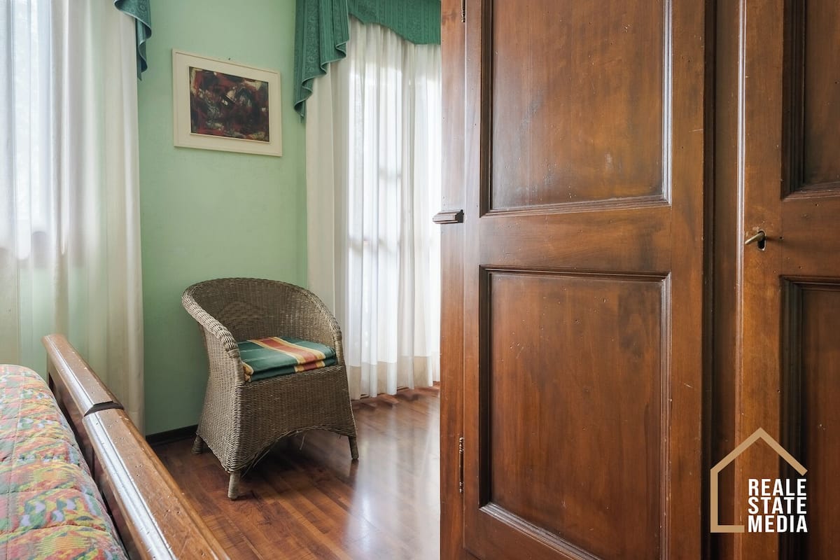 A room with a wooden door, a wicker chair, a bed with a colorful bedspread, and a painting on the wall.