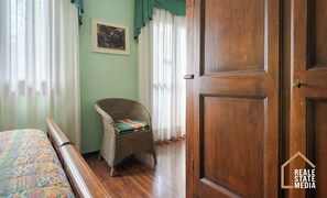 A room with a wooden door, a wicker chair, a bed with a colorful bedspread, and a painting on the wall.