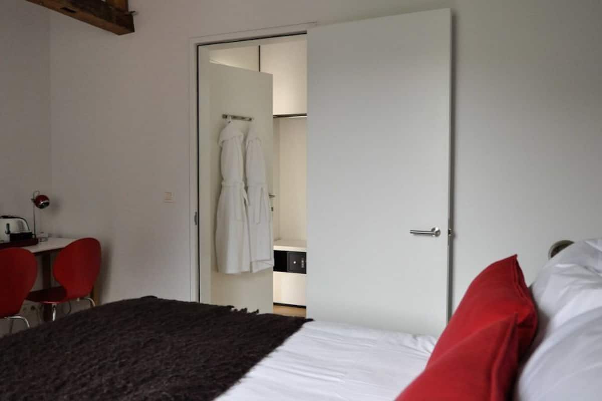 A bedroom with a bed, a desk, two chairs, a wardrobe, and a bathroom visible through an open door.