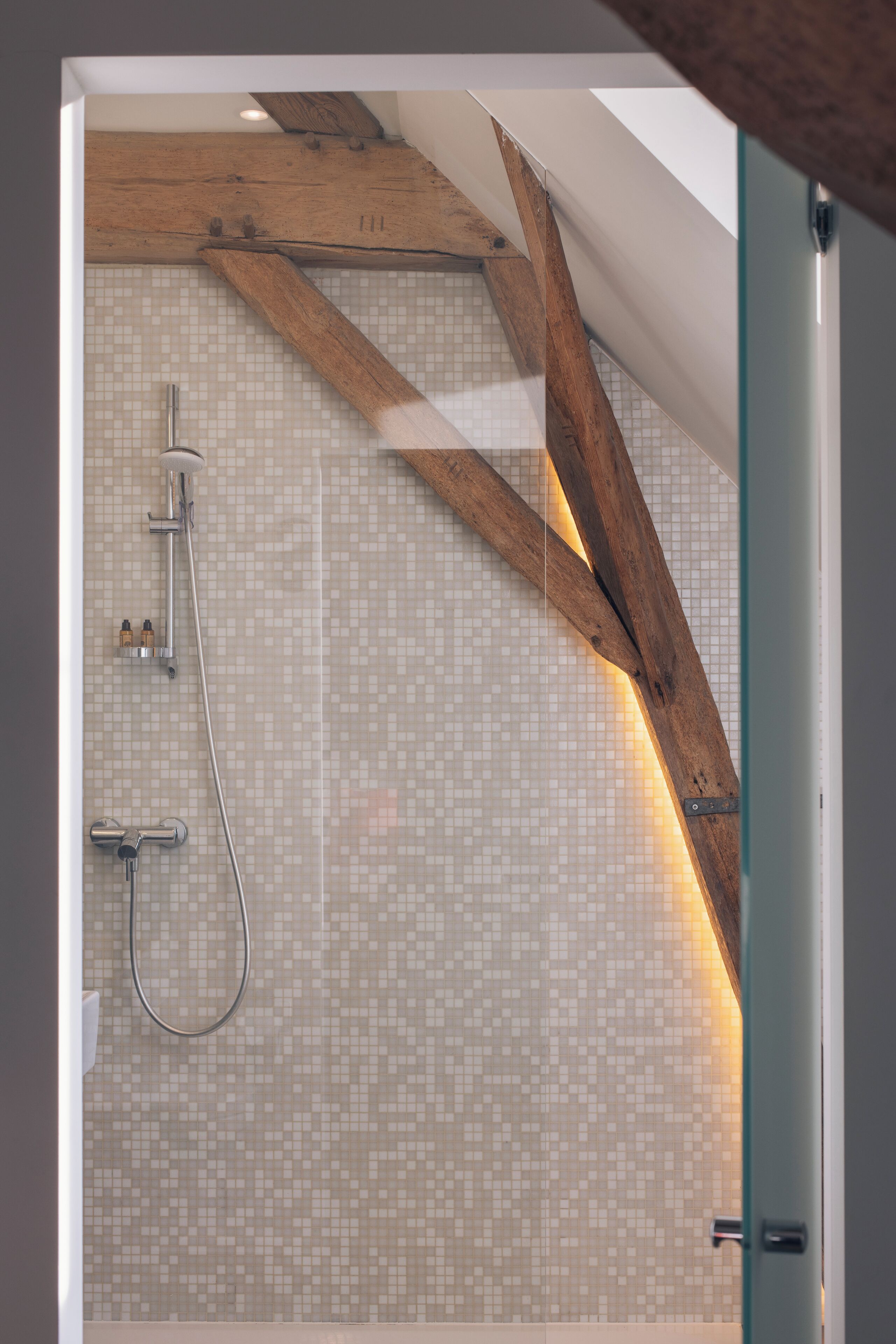 Een douche met een houten balk in het plafond, betegelde wanden en een glazen deur.