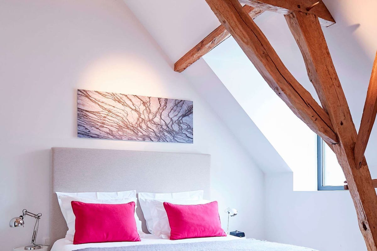 A bedroom with a sloped ceiling, wooden beams, a bed with white and pink bedding, a gray headboard, and a marble artwork above the bed.
