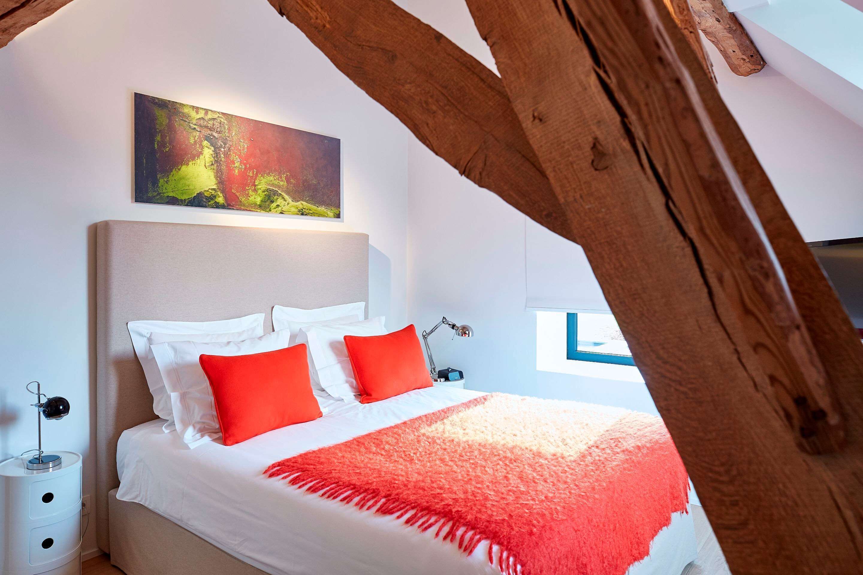 Une chambre à coucher avec un lit, du linge de lit blanc, des oreillers rouges, un plaid rouge, une table de chevet avec une lampe et un tableau accroché au mur.