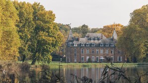 A large, historic building with multiple windows and a prominent tower, surrounded by trees and a body of water. - Park 7 (Huldenberg)