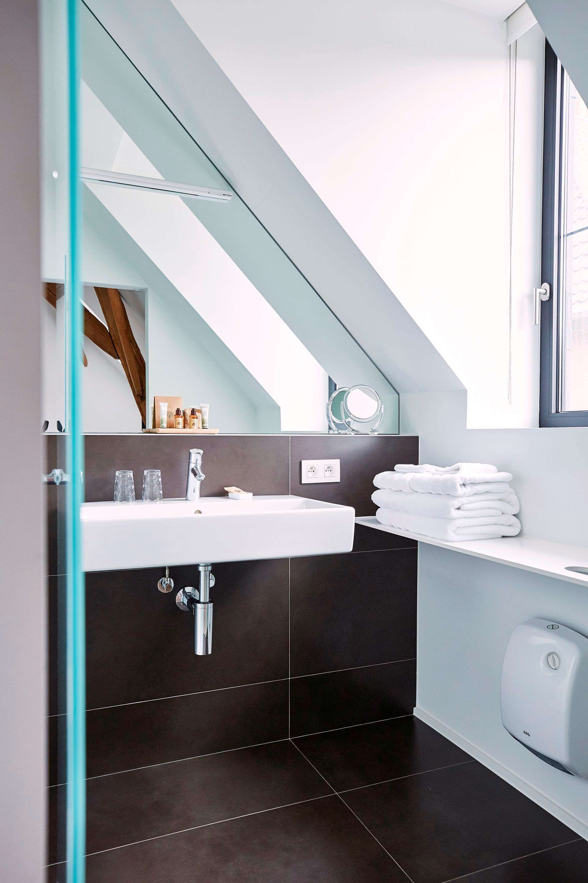 Een moderne badkamer met een douchecabine van glas, een witte wastafel, donker betegelde wanden en een raam met uitzicht.